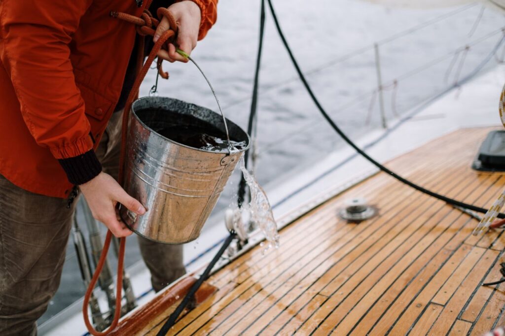 Close-up of a man washing a sailboat deck with water from a metal bucket outdoors.